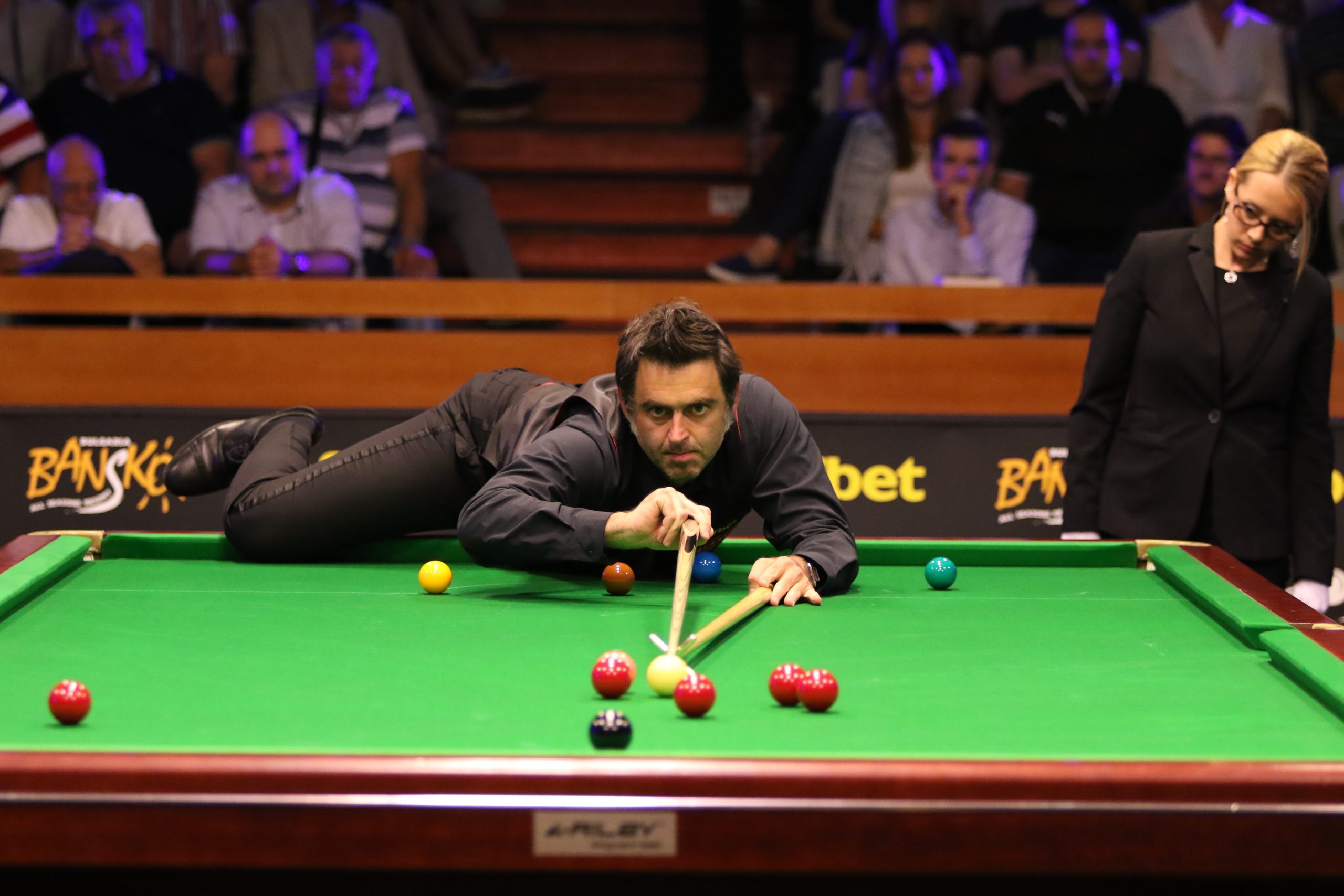 Middle aged white male snooker player leaning over table, with right leg raised and resting on the edge, preparing to take a shot