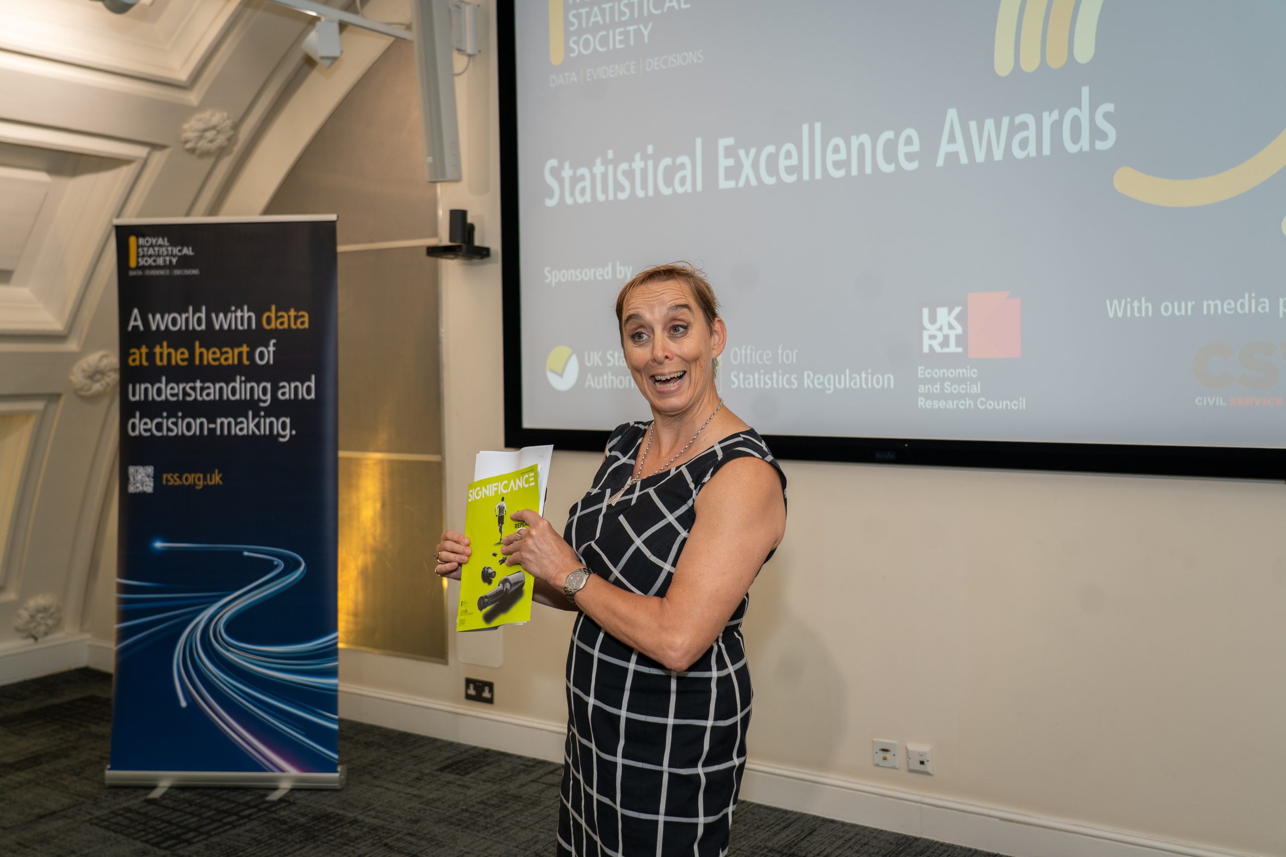 Woman smiling and holding magazine in front of a large screen reading 'Statistical Excellence Awards'