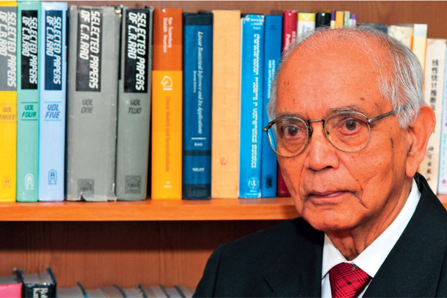 Elderly Indian man wearing glasses and standing in front of a bookcase containing his works
